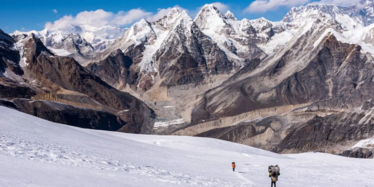 Trekkers and porters carrying heavy loads across a vast, snow-covered glacier pass toward rugged, jagged Himalayan peaks.