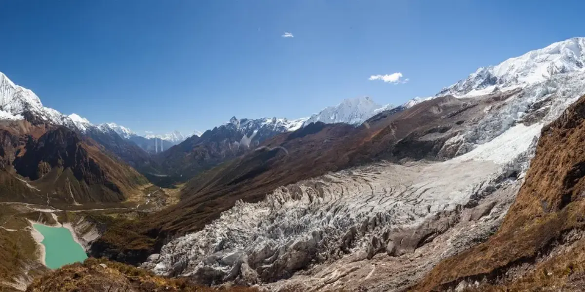 Panoramic view of a vast sprawling glacier tongue, turquoise glacial lake, and surrounding snow-capped Himalayan peaks beneath a blue sky.