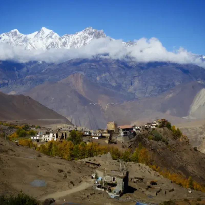 Elevated view of an ancient cliffside mountain village in an arid, high-altitude valley beneath a massive snow-capped range.