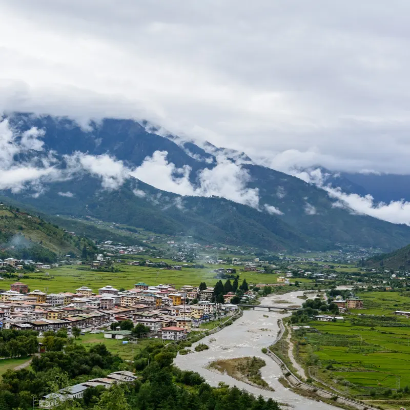 Panoramic view of Paro Valley town and river winding past green rice terraces beneath misty mountains in Bhutan.