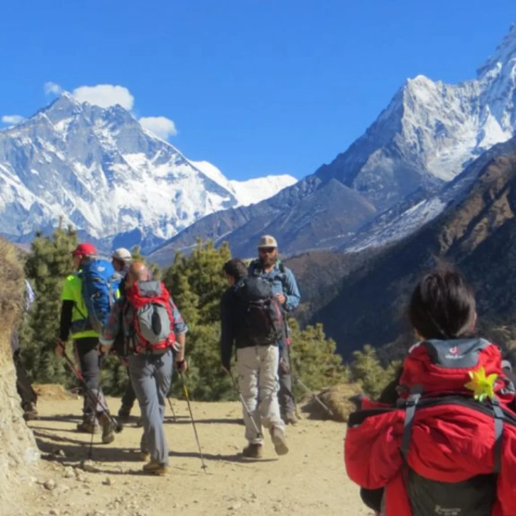 A group of trekkers with backpacks walking along a high dirt mountain trail in the Khumbu region toward the snow-capped summit of Mount Everest.