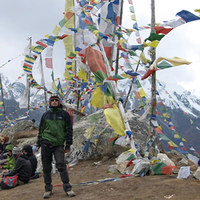 Trekker in outdoor gear standing beside towering prayer flags at a windy Himalayan mountain pass with snowy peaks in the background.