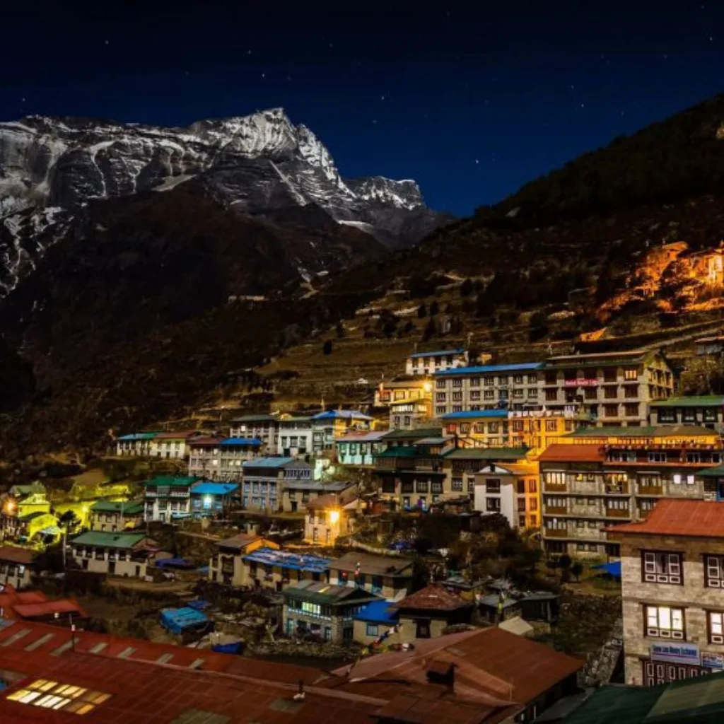 Night view of the terraced Sherpa town of Namche Bazaar illuminated by warm lights beneath a dark starry sky and snow-dusted Himalayan peaks.