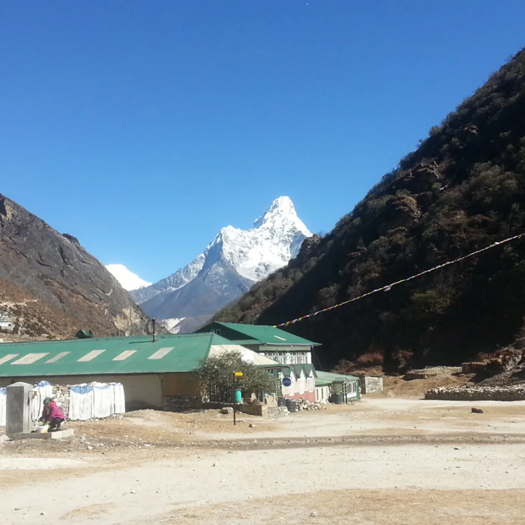 Green-roofed teahouse lodge and courtyard in the Khumbu Valley with the sharp, snow-covered summit of Mount Ama Dablam rising under a clear blue sky.