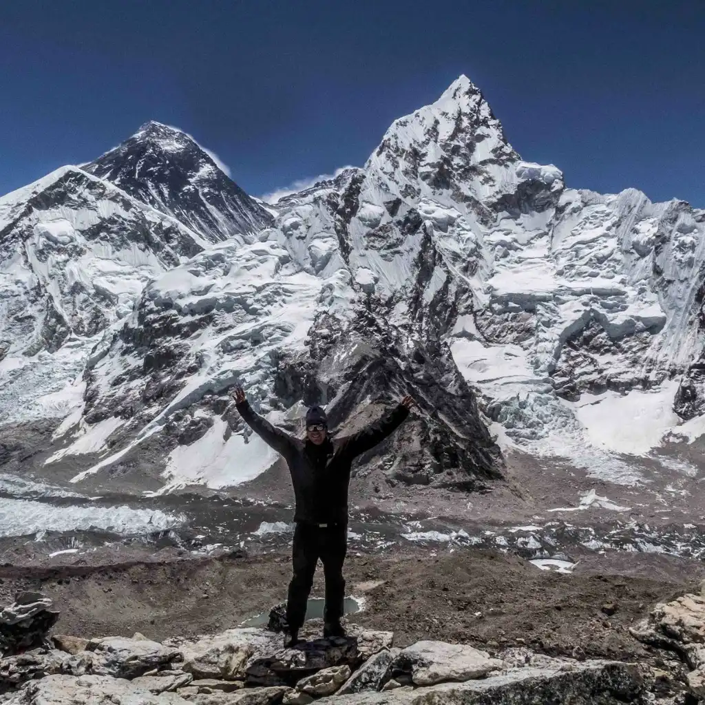 A male trekker standing on a rocky outcrop with arms raised in celebration, with Mount Everest and Mount Nuptse towering directly behind him.