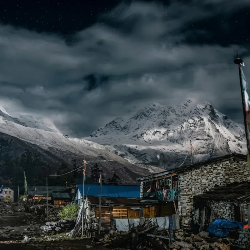 Traditional stone lodges under a dramatic night sky with moonlit clouds drifting across a massive, snow-covered mountain face.