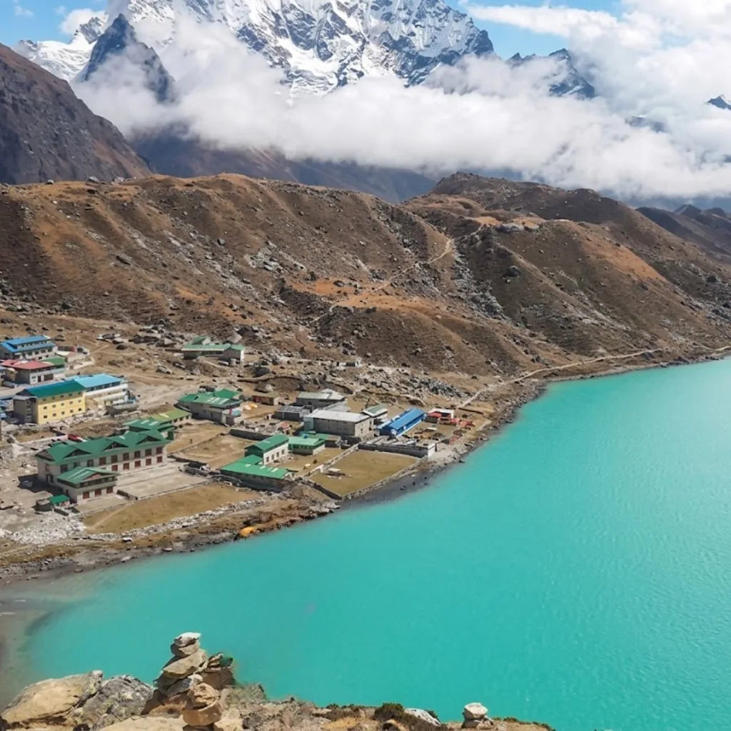Elevated view of the turquoise waters of Gokyo Lake with lakeside stone lodges beneath rolling mountain ridges and snow-capped summits in Nepal.