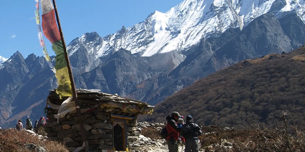 Small rustic stone shrine with Buddhist prayer flags overlooking massive snow-capped Himalayan mountain peaks.