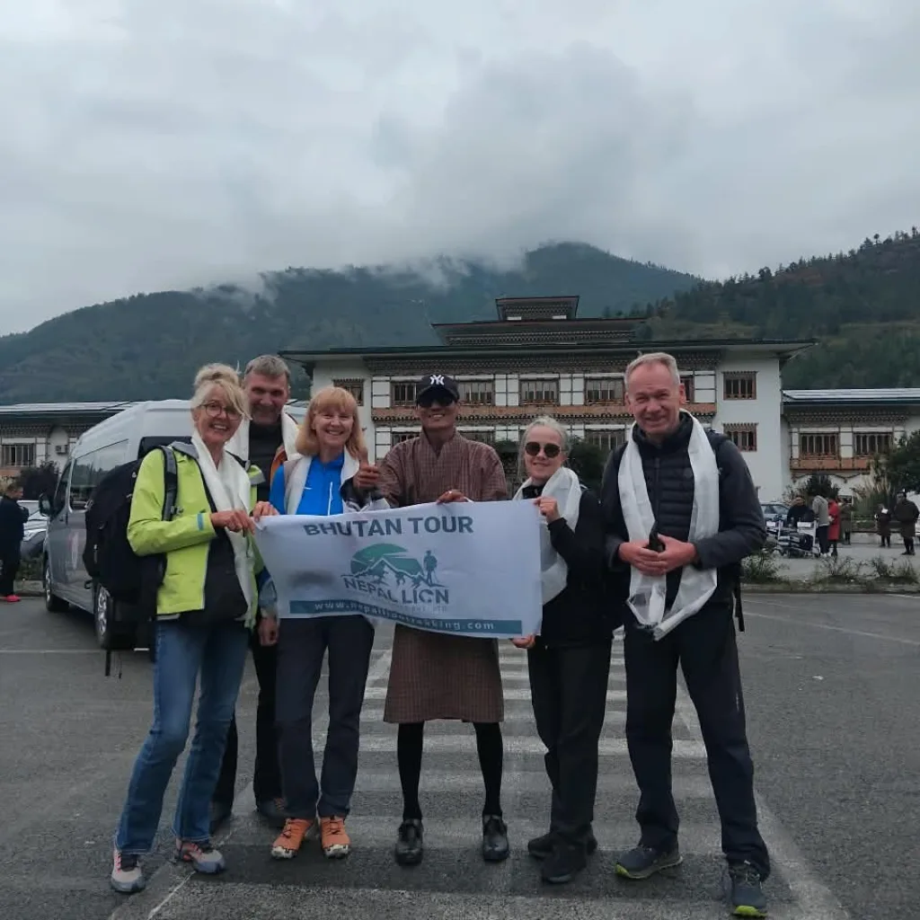 Tour group holding a "Bhutan Tour" banner with a local guide in traditional dress in front of a Bhutanese building.