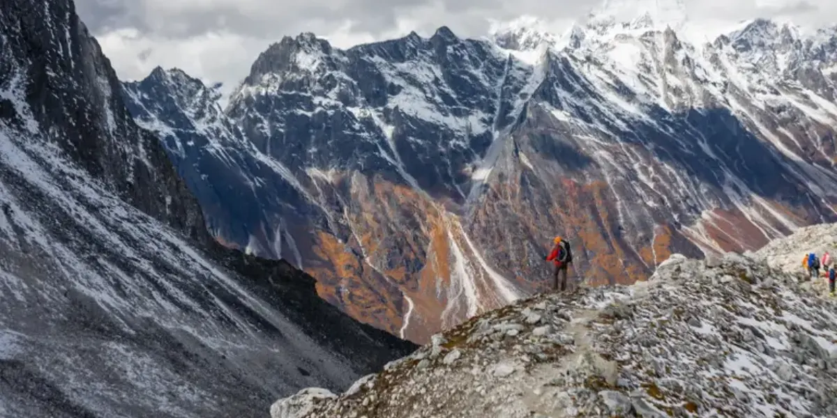 Trekker in an orange jacket walking along a rocky, snow-dusted ridge trail with sheer, jagged Himalayan mountain walls in the background.